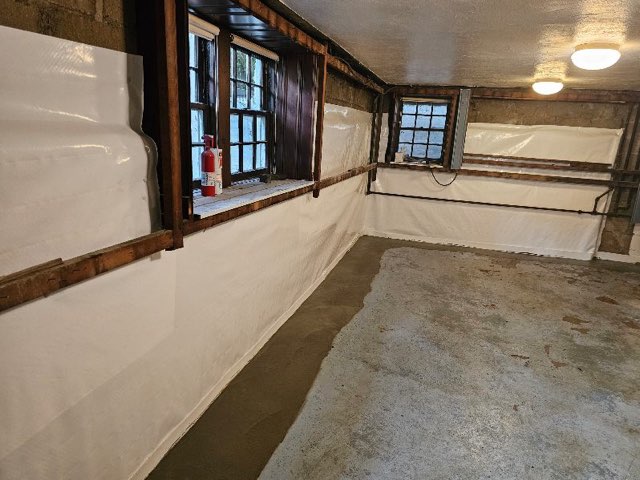 Basement with white waterproof lining on walls, concrete floors, and a window letting in natural light. A fire extinguisher is on the window ledge.