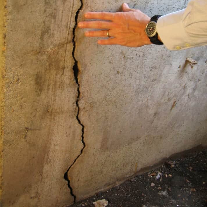 A hand touches a rough, cracked concrete wall, showing a large vertical fracture. The setting is informal, conveying concern and inspection.