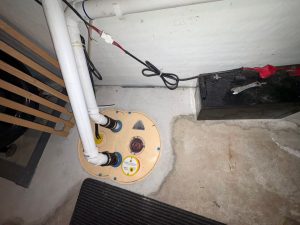 Basement corner with a sump pump and piping, adjacent to a black battery. Gray floor and white walls create a utilitarian, functional feel.
