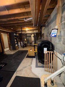 A dimly lit basement with exposed wood beams and concrete walls. It contains a furnace, water heater, shelves with paint cans, and some plumbing. The atmosphere is utilitarian.