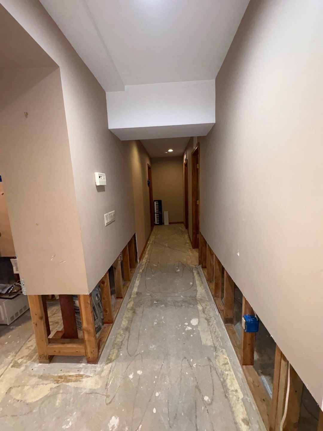 Long hallway under renovation with bare drywall and exposed wood framing at the bottom. The floor is stripped, showing concrete and some wiring.
