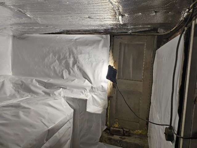 Dimly lit basement room with white plastic-covered walls and ceiling. A closed wooden door is illuminated by a single light, creating a stark contrast.