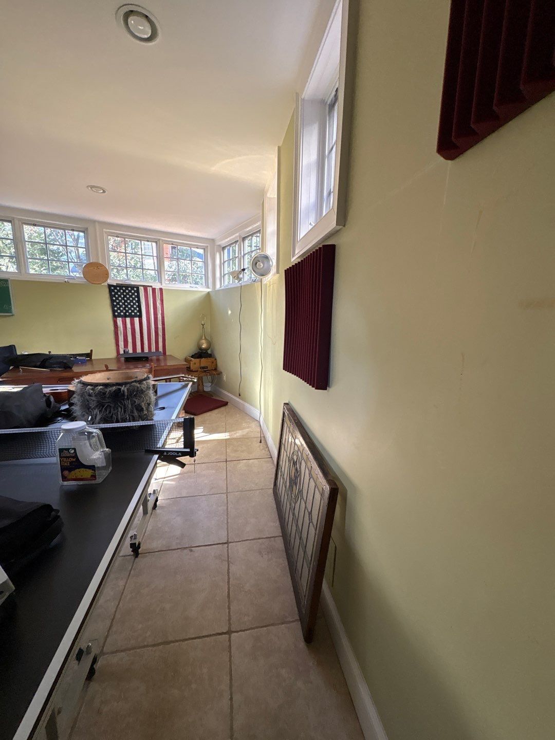 A bright home office with green walls and an American flag hanging. Sunlight streams through large windows. Items are scattered, creating a cozy yet cluttered feel.