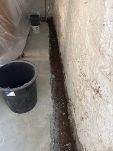 A black bucket sits on a concrete floor, near a narrow trench alongside a white-plastered wall. The scene suggests ongoing construction work.