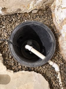 A black sump pump basin with a white PVC pipe inside, surrounded by gravel and concrete. The setting appears to be a basement or utility area.